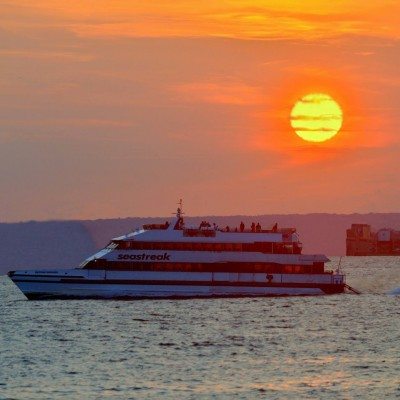 A Seastreak ferry cruises through the harbor with a perfect sunset on the horizon.